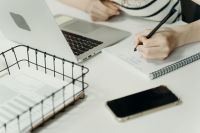Sales Manager aan bureau, schrijvend in schrift bij laptop en telefoon.