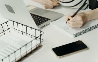 Sales Manager aan bureau, schrijvend in schrift bij laptop en telefoon.